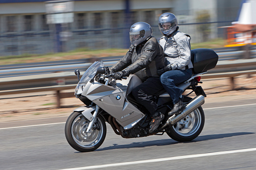 Motorcyclist and a passenger on a silver sport bike traveling on a roadway.