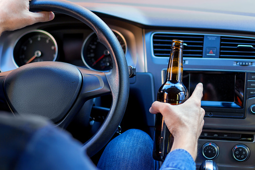 Closeup male hand is holding beer behind steering wheel of car.