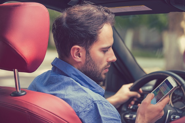Man in a blue shirt looking down at his smartphone while driving, with one hand on the steering wheel and the other holding the phone.