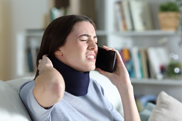 Woman wearing a neck brace grimacing in pain while talking on the phone in a home setting.