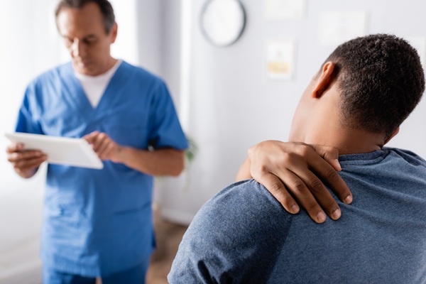 A man clutching his neck and shoulder while a healthcare professional in blue scrubs reviews information on a tablet in the background.