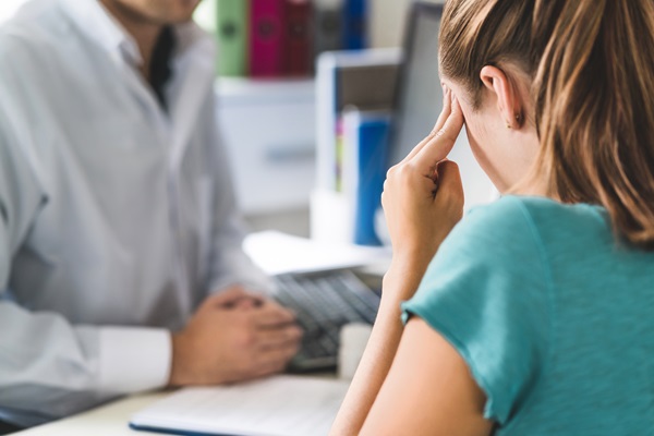 An over-the-shoulder view of a woman sitting in a clinical setting, pressing her fingers to her temple in distress while speaking with a brain injury doctor in New Mexico about her symptoms.