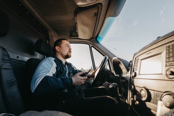 Commercial van driver holding a smartphone while seated behind the steering wheel, looking away from the road, distracted driving safety concern